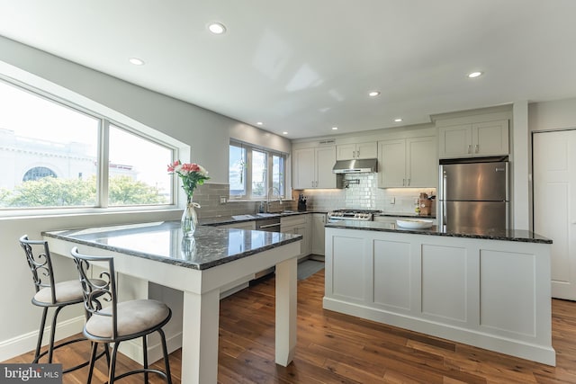 kitchen with appliances with stainless steel finishes, a kitchen bar, backsplash, dark stone counters, and dark wood-type flooring