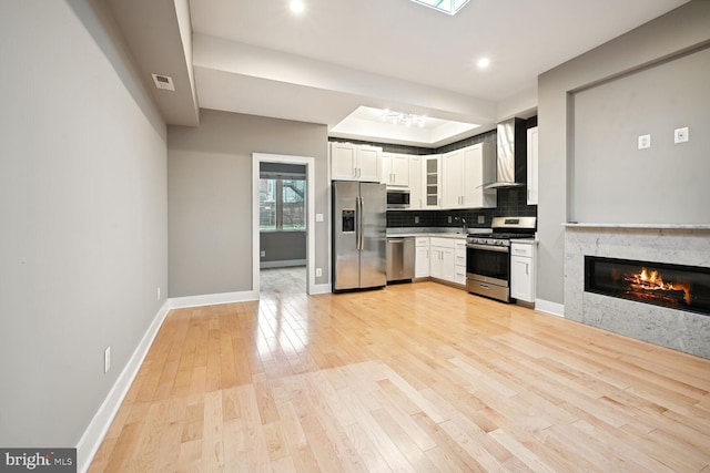 kitchen with white cabinets, a premium fireplace, wall chimney range hood, appliances with stainless steel finishes, and light wood-type flooring