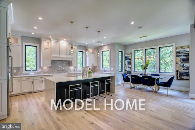 kitchen featuring light wood-type flooring, a kitchen island with sink, white cabinets, hanging light fixtures, and decorative backsplash