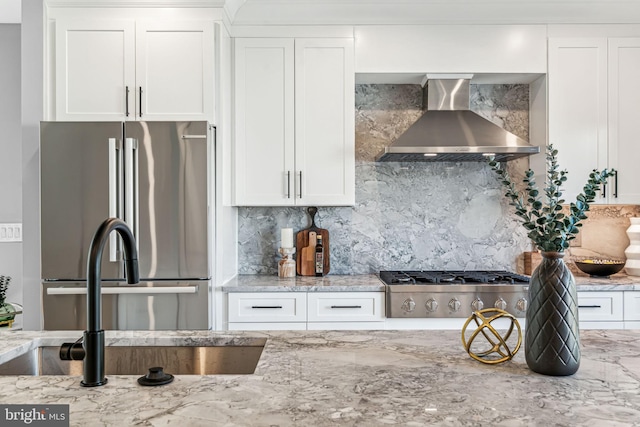 kitchen featuring appliances with stainless steel finishes, white cabinetry, light stone counters, tasteful backsplash, and wall chimney range hood