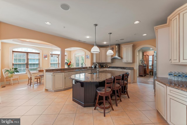 kitchen featuring an island with sink, pendant lighting, dark stone counters, cream cabinets, and wall chimney range hood