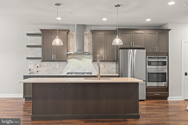 kitchen with a kitchen island with sink, dark wood-type flooring, hanging light fixtures, wall chimney exhaust hood, and stainless steel appliances