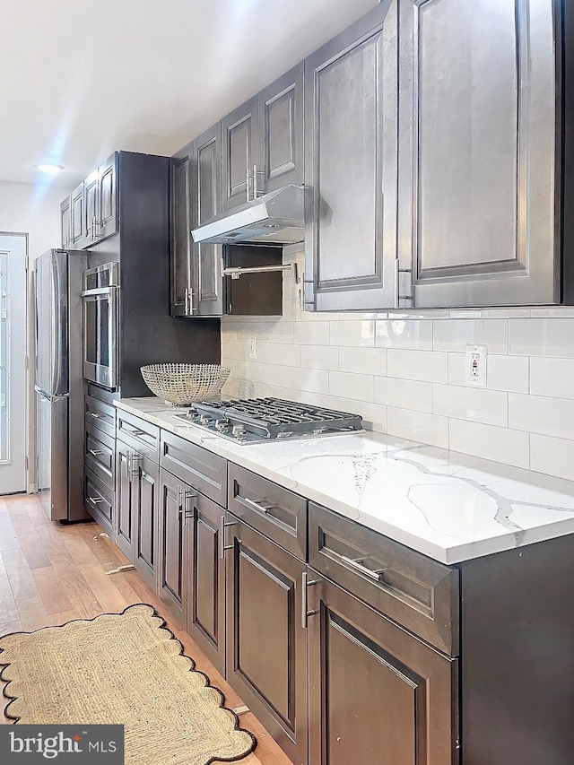 kitchen with appliances with stainless steel finishes, light wood-type flooring, light stone counters, and backsplash