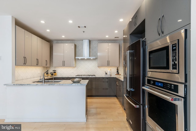 kitchen with gray cabinetry, appliances with stainless steel finishes, a sink, a peninsula, and wall chimney exhaust hood