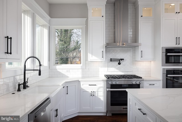 kitchen featuring white cabinets, wall chimney range hood, appliances with stainless steel finishes, light stone countertops, and decorative backsplash