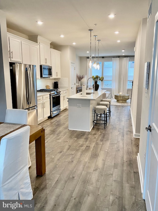 kitchen with light hardwood / wood-style floors, an island with sink, white cabinetry, appliances with stainless steel finishes, and decorative light fixtures