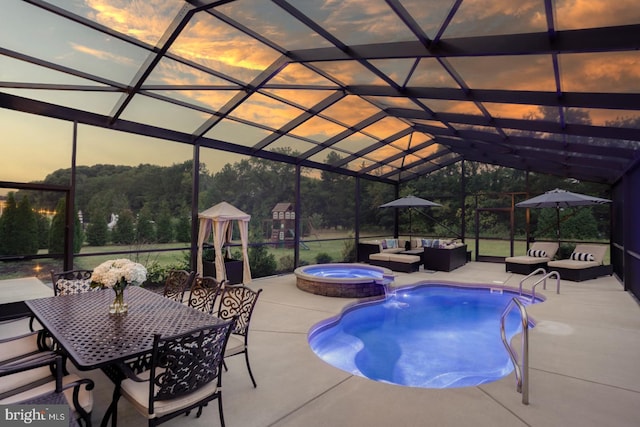 pool at dusk featuring glass enclosure, an in ground hot tub, and a patio