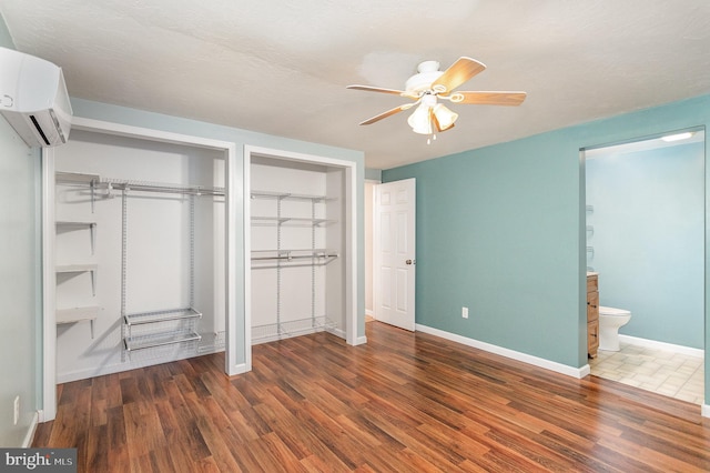 unfurnished bedroom with a wall unit AC, dark hardwood / wood-style floors, multiple closets, ensuite bathroom, and ceiling fan