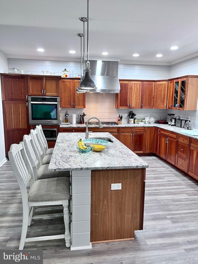 kitchen with an island with sink, light wood-type flooring, exhaust hood, and decorative light fixtures