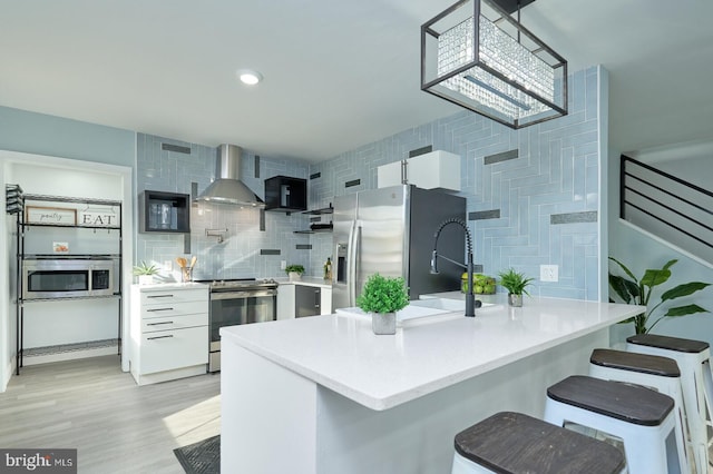 kitchen featuring white cabinets, wall chimney range hood, appliances with stainless steel finishes, light wood-type flooring, and decorative backsplash