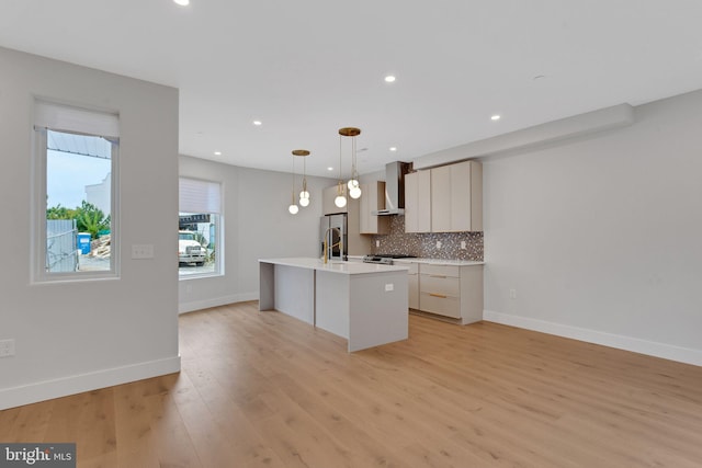 kitchen featuring wall chimney exhaust hood, an island with sink, pendant lighting, light wood-type flooring, and tasteful backsplash