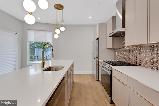 kitchen with wall chimney range hood, appliances with stainless steel finishes, sink, hanging light fixtures, and light hardwood / wood-style floors