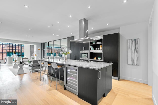 kitchen with stainless steel oven, wine cooler, light hardwood / wood-style flooring, island exhaust hood, and a center island with sink