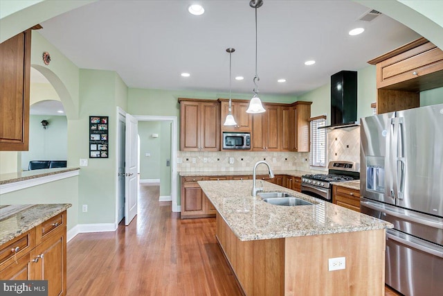 kitchen featuring appliances with stainless steel finishes, an island with sink, light stone counters, light hardwood / wood-style flooring, and range hood