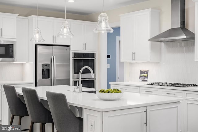 kitchen featuring white cabinets, a center island with sink, wall chimney range hood, appliances with stainless steel finishes, and a breakfast bar