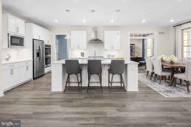 kitchen featuring a kitchen island with sink, white cabinets, hanging light fixtures, wall chimney range hood, and stainless steel appliances