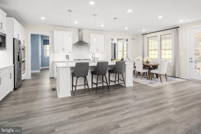 kitchen with white cabinets, wall chimney exhaust hood, pendant lighting, wood-type flooring, and a center island with sink