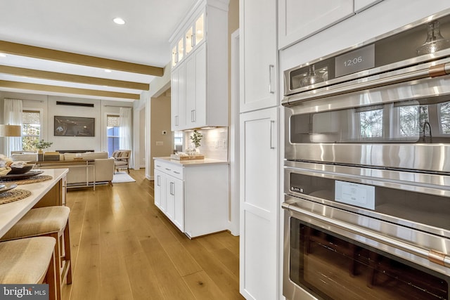 kitchen featuring tasteful backsplash, white cabinets, stainless steel double oven, beam ceiling, and light hardwood / wood-style flooring