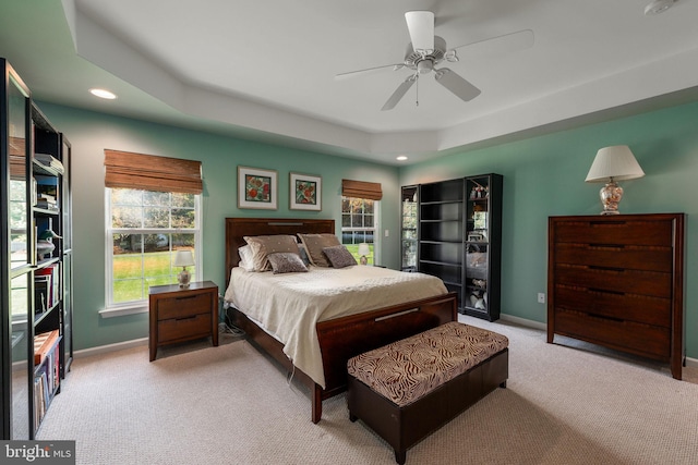 bedroom featuring a raised ceiling, multiple windows, light colored carpet, and ceiling fan