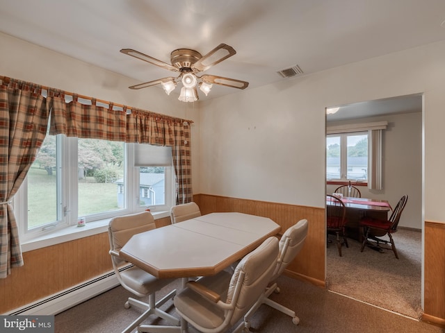 carpeted dining space with ceiling fan, wooden walls, baseboard heating, and a wealth of natural light