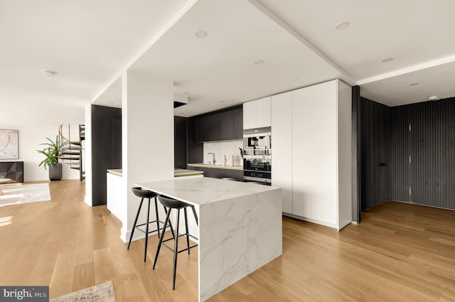 kitchen with light wood-type flooring, a kitchen breakfast bar, decorative backsplash, light stone countertops, and white cabinets