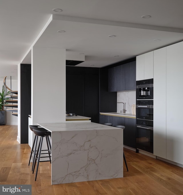 kitchen with decorative backsplash, a breakfast bar, black double oven, light hardwood / wood-style floors, and white cabinetry