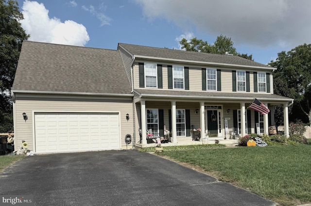 colonial-style house featuring aphalt driveway, an attached garage, covered porch, a shingled roof, and a front yard