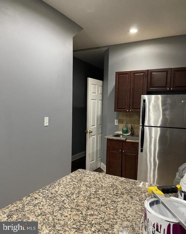 kitchen with dark brown cabinets, light stone counters, sink, backsplash, and stainless steel fridge