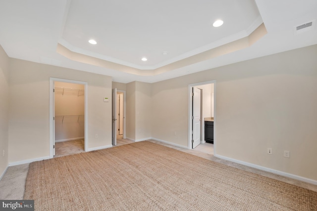 unfurnished bedroom featuring light colored carpet, a raised ceiling, ensuite bath, and a walk in closet