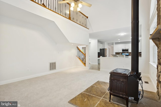 carpeted living room with stairway, a ceiling fan, a wood stove, visible vents, and a towering ceiling