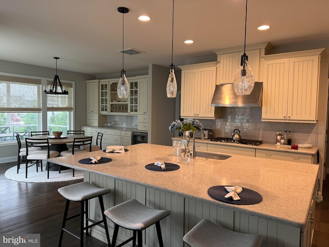 kitchen with a kitchen island with sink, dark hardwood / wood-style floors, tasteful backsplash, wall chimney exhaust hood, and stainless steel gas cooktop