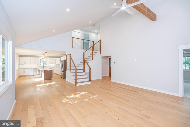 unfurnished living room with beam ceiling, ceiling fan with notable chandelier, light wood-type flooring, and high vaulted ceiling