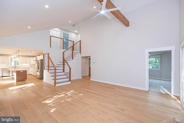 unfurnished living room featuring sink, ceiling fan with notable chandelier, light hardwood / wood-style floors, beamed ceiling, and high vaulted ceiling