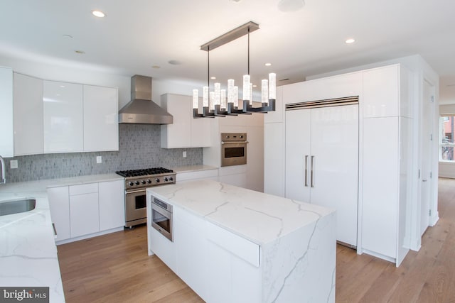 kitchen with light hardwood / wood-style floors, white cabinetry, built in appliances, pendant lighting, and wall chimney range hood