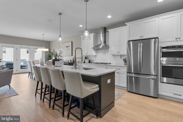 kitchen with white cabinets, hanging light fixtures, wall chimney range hood, a center island with sink, and appliances with stainless steel finishes