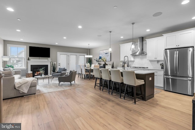 kitchen featuring white cabinets, stainless steel appliances, light wood-type flooring, a center island with sink, and wall chimney range hood