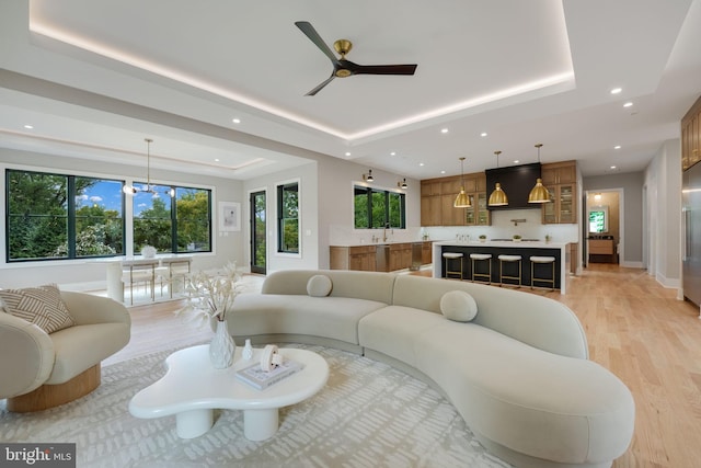 living room with ceiling fan with notable chandelier, light hardwood / wood-style floors, a tray ceiling, and sink