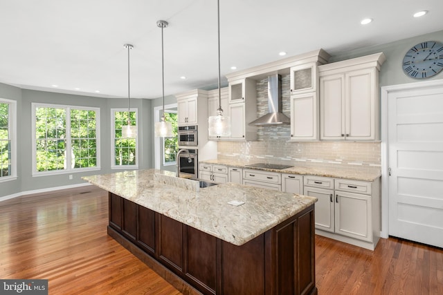 kitchen with dark wood-type flooring, a large island with sink, wall chimney range hood, hanging light fixtures, and white cabinetry