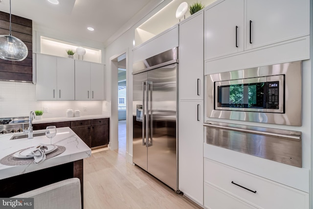 kitchen with light wood-type flooring, backsplash, built in appliances, decorative light fixtures, and white cabinetry