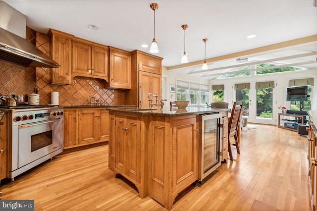 kitchen with wine cooler, decorative light fixtures, a center island with sink, wall chimney exhaust hood, and designer stove