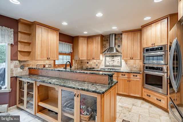 kitchen with sink, wall chimney exhaust hood, backsplash, stainless steel appliances, and dark stone counters