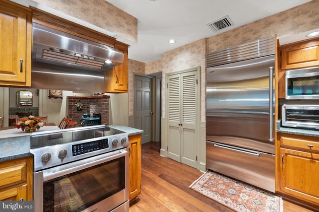 kitchen with premium appliances, dark stone counters, exhaust hood, and light wood-type flooring