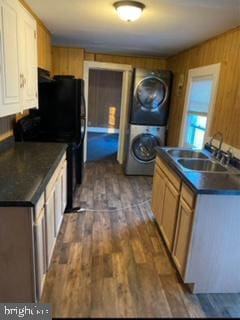 kitchen featuring dark hardwood / wood-style floors, sink, stacked washing maching and dryer, wooden walls, and black refrigerator