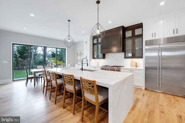 kitchen featuring light wood-type flooring, a kitchen island with sink, hanging light fixtures, stainless steel appliances, and light stone countertops