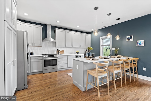 kitchen with wall chimney range hood, appliances with stainless steel finishes, hardwood / wood-style floors, hanging light fixtures, and white cabinets