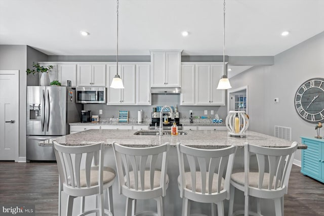 kitchen featuring dark wood-type flooring, light stone counters, stainless steel appliances, decorative light fixtures, and a center island with sink