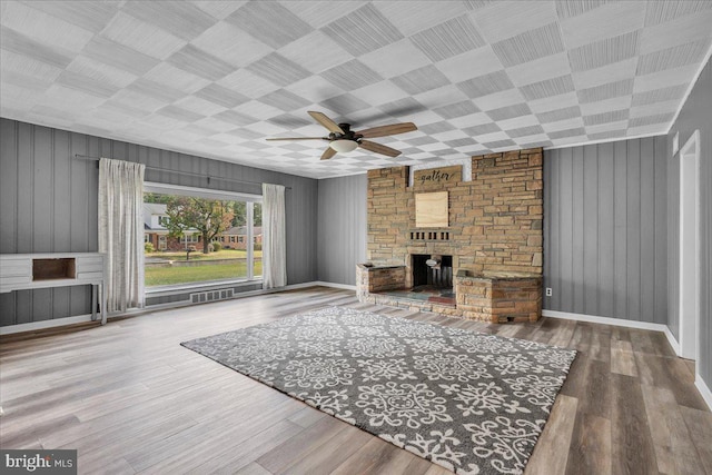 unfurnished living room featuring a stone fireplace, wooden walls, wood-type flooring, and ceiling fan