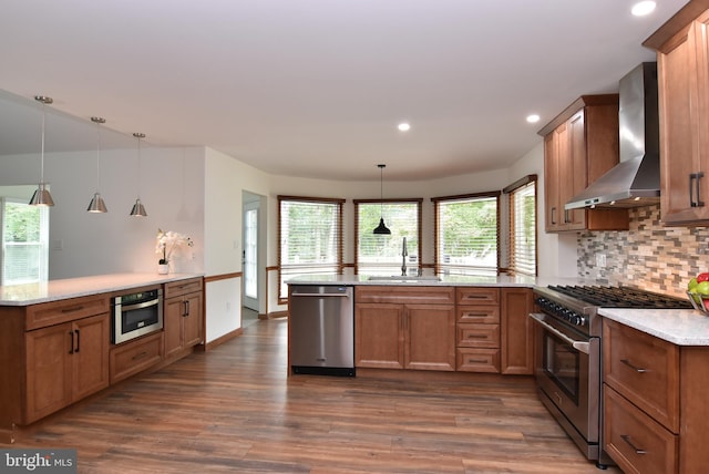 kitchen with kitchen peninsula, hanging light fixtures, appliances with stainless steel finishes, wall chimney exhaust hood, and sink