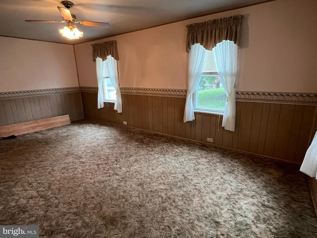 empty room with a wealth of natural light, dark colored carpet, ceiling fan, and wooden walls