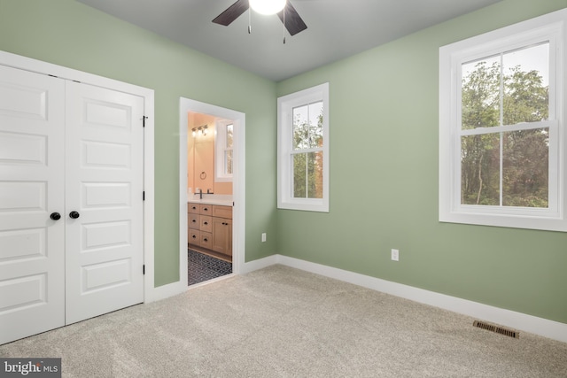 unfurnished bedroom featuring carpet, ensuite bath, multiple windows, and ceiling fan